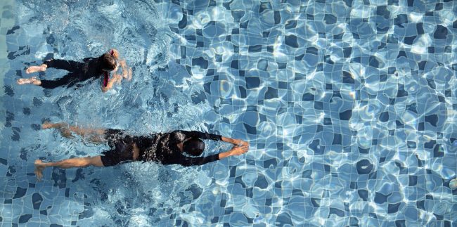 Two boys funny swimming together in clear water pool, taken from aerial view.